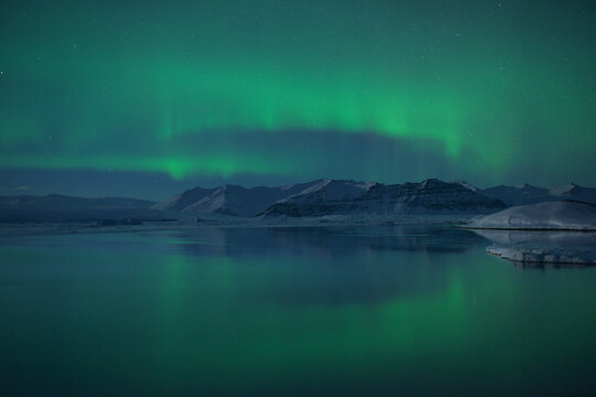 View of the ethereal green aurora borealis dances above the icy lagoon, reflecting in the still waters beneath the snow-capped mountains, Jokulsarlon, Sveitarfélagio Hornafjorour, Iceland.