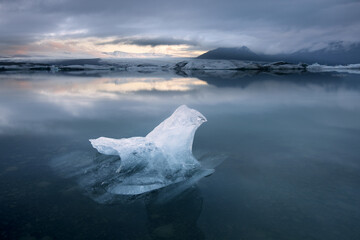 View of a pristine, solitary ice floe adrift in the tranquil, mirrored waters beneath a brooding sky, reflecting the distant mountains, Jokulsarlon, Sveitarfélagio Hornafjorour, Iceland.