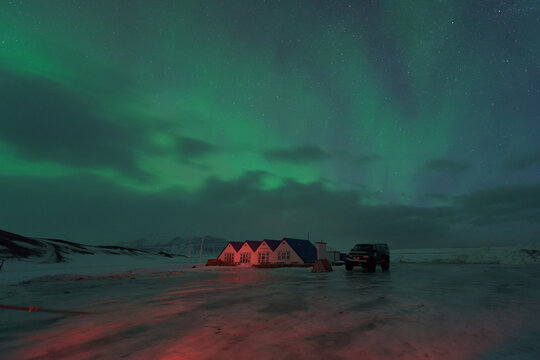 View of the aurora borealis painting the sky green above a row of houses and a black vehicle in a cold, snowy landscape, Jokulsarlon, Sveitarf&eacute;lagio Hornafjorour, Iceland.