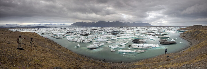 View of milky blue waters dotted with luminous icebergs under a cloudy sky, photographers capturing the stark beauty, Jokulsarlon, SveitarfÃ©lagiÃ° HornafjÃ¶rÃ°ur, Iceland.