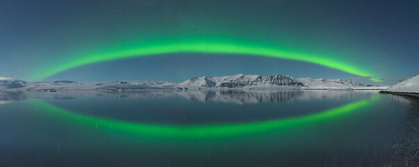 View of a vibrant green aurora borealis arcs across the night sky, reflected in the still waters below, illuminating the snow-covered mountains, Jokulsarlon, Sveitarfélagið Hornafjörður, Iceland.
