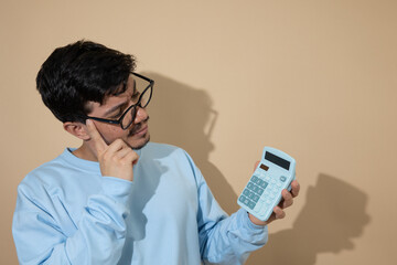 Man with glasses looking at a blue calculator with a thoughtful expression on a light brown background with copy space, concept of financial analysis and economic planning