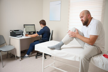 Fototapeta premium Male patient being examined in clinic for knee pain. Male suffering from knee pain osteoarthritis. Close up man holding his knee due to physical injury while sitting in room.