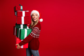 Funny joyful young woman in a Santa hat carries a tower of Christmas gifts against a bright red...