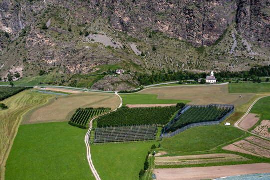 Aerial view of neatly patterned fields and orchards nestled beneath rugged mountains, a church standing sentinel over the tranquil scene, Molini di Tures, Trentino-Alto Adige/South Tyrol, Italy.