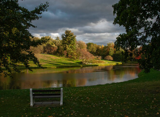 View of a bench sits overlooking a tranquil lake reflecting the vibrant autumn foliage under a dramatic, cloudy sky, Poughkeepsie, New York, United States.