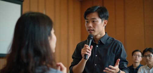 Young Asian man with microphone gives presentation. People listen attentively in classroom setting. Focus on public speaking skills development and practice.