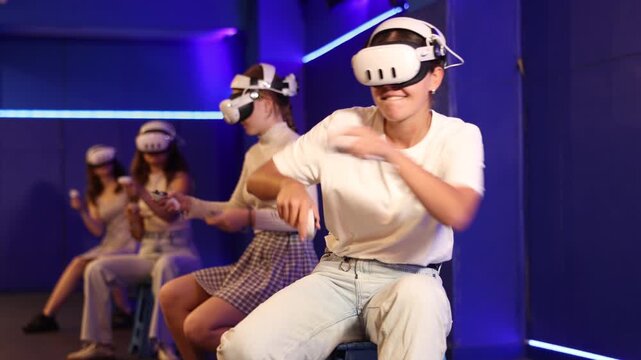 Group of excited young girls sitting with handheld controllers in hands engaged in virtual world experiencing through VR glasses covering face. Concept of immersion and interactivity in game