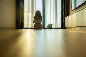 Confident woman using laptop and planning in new home sitting on floor. Bright sunlight shines through transparent silk curtain hanging at room. Ray of sun illuminate apartment at sunset or sunrise.