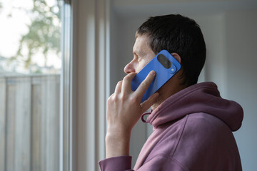 Young man with dark hair, wearing a pink hoodie, is engaged in a phone conversation while standing by a window, showcasing a moment of connection and communication