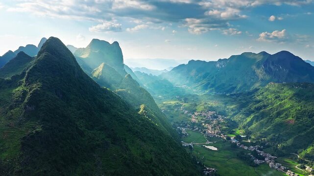 Aerial shot of a spectacular green mountain valley with sun rays breaking through clouds over a small village in a rural area. 