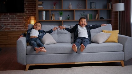 Happy African American little boys jumping on couch joyful positive ethnic children carefree active siblings sons kids running in living room jump on comfy sofa playing family having fun joy indoors