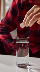 Senior hand holding round tablet above glass of water before dissolving. Concept of pharmacy instruction, home treatment education, and health awareness.