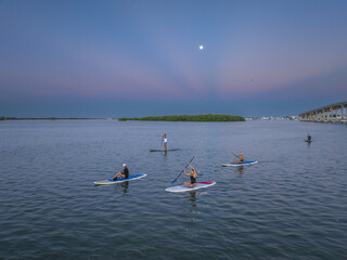Aerial view of paddleboarders gliding across the tranquil waters under a soft, moonlit sky near the distant bridge, Progreso, Yucatan, Mexico.