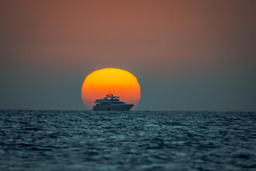 Aerial view of a yacht sailing into the sunset, its silhouette a stark contrast against the fiery orb, as the deep blue sea reflects the sky's warm hues, Progreso, Yucatan, Mexico.