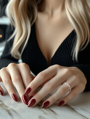 close-up of hands with long glossy dark red nails resting on marble surface, featuring delicate ring with small stones and contrasted by black ribbed deep V-neck top