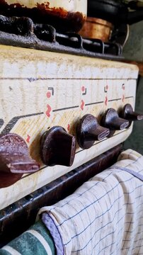 Vertical, close-up view of an old gas stove front panel with control knobs. Brown plastic knobs with red and black markings are on a cream enamel surface, with a striped kitchen towel hanging below.