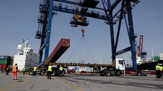 Wide-angle video shot of a bustling port, capturing workers and machinery loading a shipping container onto a truck under a clear blue sky.