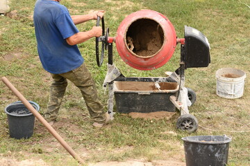 A man in a blue T-shirt and green pants pulls the round handle of a red concrete mixer. Two black buckets and one white bucket are on the grass nearby. Rural construction and manual labor scene.