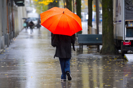 Rear view of young woman with orange umbrella walking on pavement of famous shopping mile named Bahnhofstrasse on a rainy autumn day. Photo taken October 27th, 2025, Zurich, Switzerland.