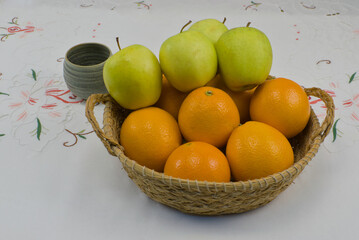 Cesta de esparto con naranjas y manzanas en su interior junto a un vaso de cerámica sobre un mantel bordado