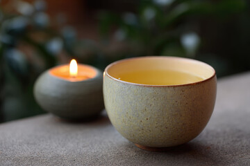 Cup of tea with warm yellow hue sits beside flickering candle, creating peaceful and cozy atmosphere, perfect for relaxation and mindfulness in tranquil setting