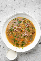 Hearty bowl of beef, vegetable, and barley soup, garnished with fresh parsley, served on a gray stone surface. A side dish of sour cream is next to it.