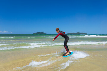 Young boy skimboarding in shallow water on a sunny beach day. Wearing wetsuit, balancing on board with determination and focus. Perfect image for themes of childhood adventure, active lifestyle, water