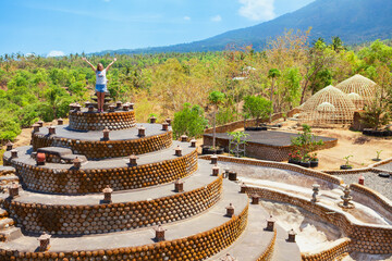 Young joyful woman stands with raised hands on buddhist stupa terrace at Aung mount slopes in Tulamben, Bali, Indonesia. Eco landmark inspired by Borobudur, surrounded by tropical forest and blue sky.
