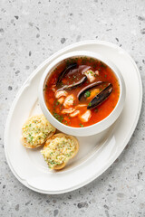 vibrant bowl of seafood soup, such as bouillabaisse or cioppino, with mussels, shrimp, and fish in a tomato-based broth. Served on a white oval plate with two slices of garlic toast 