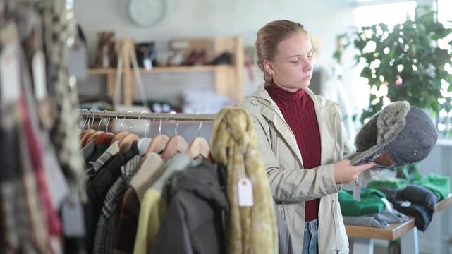 Attentive teen girl customer holding warm fur hat during winter sale in retail outlet 