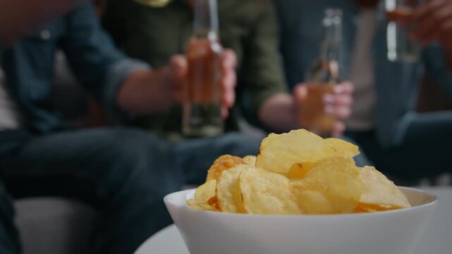 Close up plate with chips snacks four friends clinking bottles on background home party group of multiracial people men women celebrating birthday spending time weekend holding alcohol drinks on couch