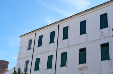 Sunlit Mediterranean-style building with white facade and teal shutters against a clear blue sky.