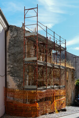 Construction scaffolding and orange safety netting surrounding a weathered stone building on a narrow street.