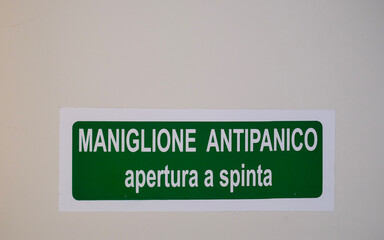 Italian emergency push-bar sign “MANIGLIONE ANTIPANICO – apertura a spinta” on a white wall.