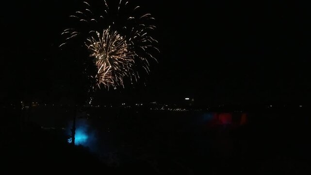 Night view of Niagara Falls as fireworks burst in the sky. The waterfall is lit red and blue through the mist, with city lights in the distance. Celebration, tourism, border pride, shared wonder.
