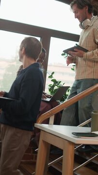 Vertical tracking shot of middle-aged Caucasian male programmers engaged in conversation while going downstairs and greeting their young Black female colleague sitting by window, giving high five