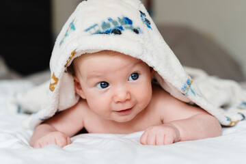 Happy curious baby child after bath with towel on head. Adorable little boy wrapped in blanket. Cutest 3 months old baby smiling in towel on bed close up at home. Child caring routine.
