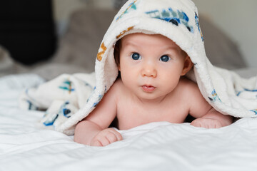 Happy curious baby child after bath with towel on head. Adorable little boy wrapped in blanket. Cutest 3 months old baby smiling in towel on bed close up at home. Child caring routine.
