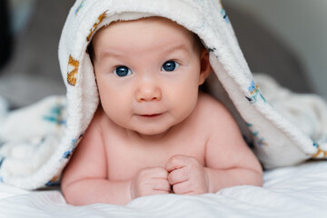 Happy curious baby child after bath with towel on head. Adorable little boy wrapped in blanket. Cutest 3 months old baby smiling in towel on bed close up at home. Child caring routine.