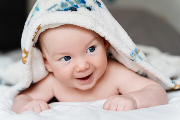 Happy curious baby child after bath with towel on head. Adorable little boy wrapped in blanket. Cutest 3 months old baby smiling in towel on bed close up at home. Child caring routine.