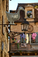 Exploring Sarlat-La-Caneda reveals a metal duck sign above a quaint cobblestone street, highlighting the allure of the decorated facades and vibrant flowers in the Dordogne valley.