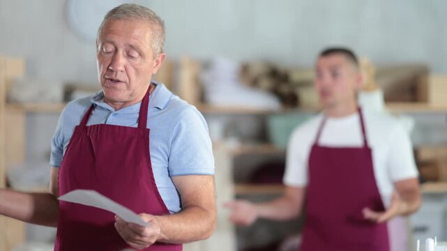  Young guy tailor scolding elderly male assistant with document for dismissal in sewing workshop 