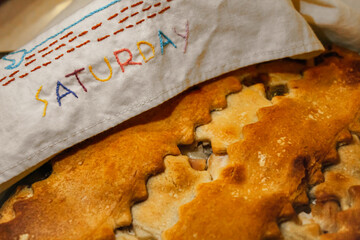 Close-up of a baked pie with a decorative lattice crust and a hand embroidered cloth that reads SATURDAY. Vintage baking background or holiday comfort food banner.