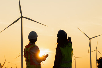 Two engineers discuss renewable energy solutions with wind turbines in the background during a sunset, promoting sustainable practices. © venusvi