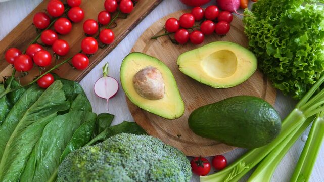 Person's hand placing half an avocado on a wooden cutting board. Surrounded by a variety of fresh organic vegetables like cherry tomatoes. Broccoli. Celery. Lettuce. And spinach for a healthy meal