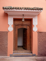 Vibrantly Peach-Walled Moroccan Building with Open Carved Wooden Door, Intricate Plasterwork, and Traditional Green Tiled Roof in Marrakech