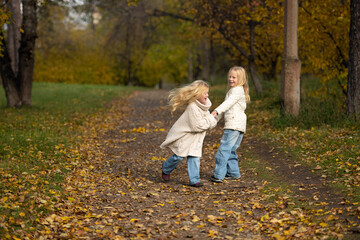 Fototapeta premium Two blonde sisters wearing a white knitted sweater and jeans, are playing in autumn park. Warm natural light and fall colours create a dreamy, emotional atmosphere of childhood and nature.