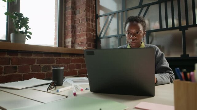 Medium shot of focused young African American female programmer or office employee in casualwear opening laptop at desk and concentrating on work