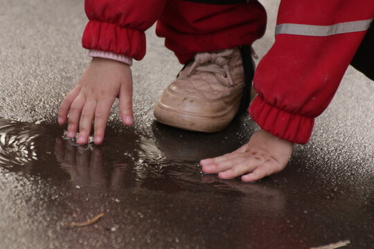  toddler in a red outdoor suit touches a puddle, gaining sensory experience. Faces not visible. Perfect for parenting, early childhood development, sensory play, and outdoor exploration content